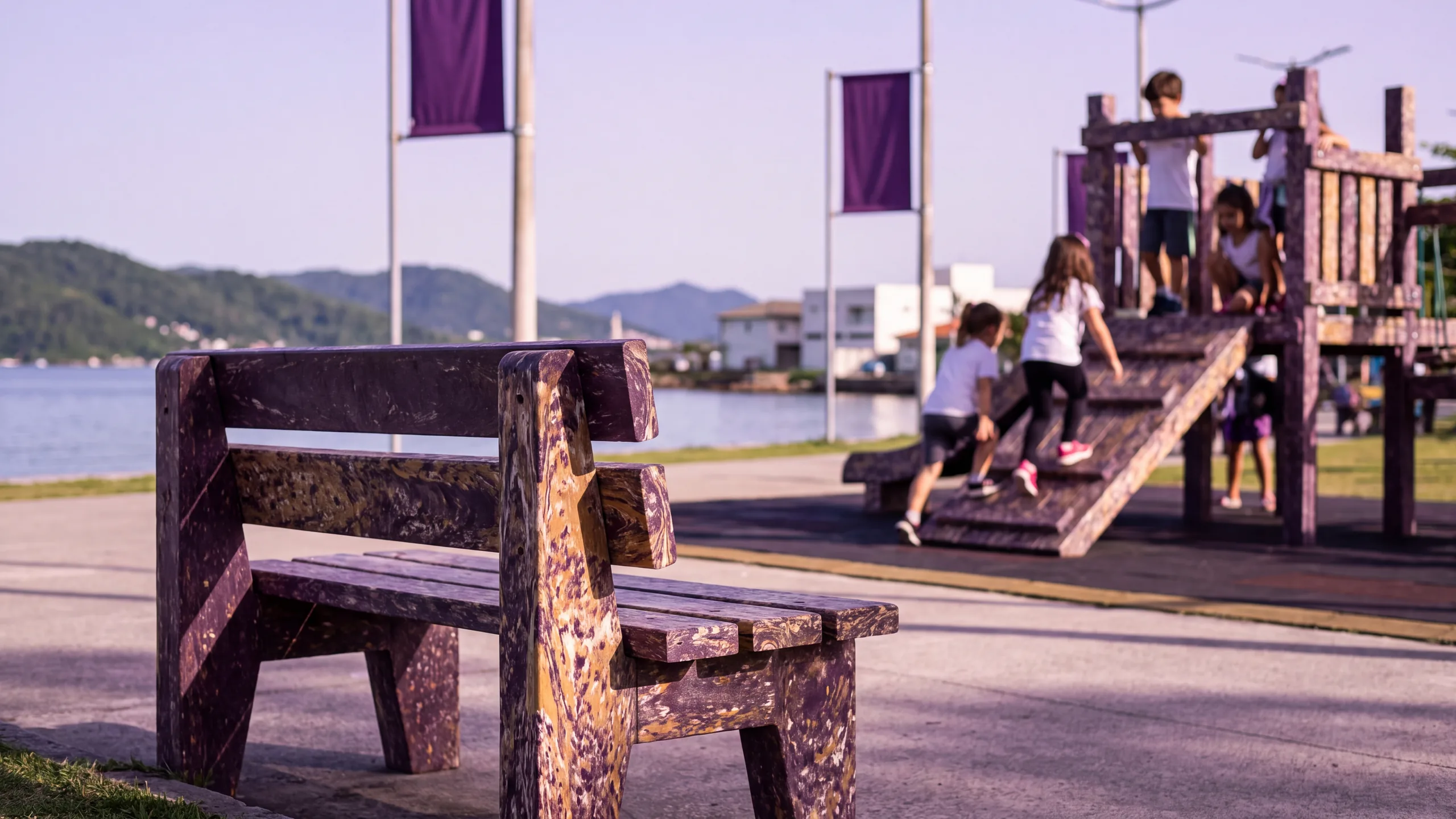 Banco de praça feito de plástico reciclado à beira da baía, com crianças brincando num brinquedo também de plástico reciclado ao fundo