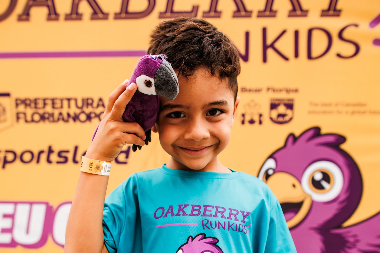 Menino de camiseta Oakberry Run Kids segura a arara de pelúcia diante do painel da corrida infantil