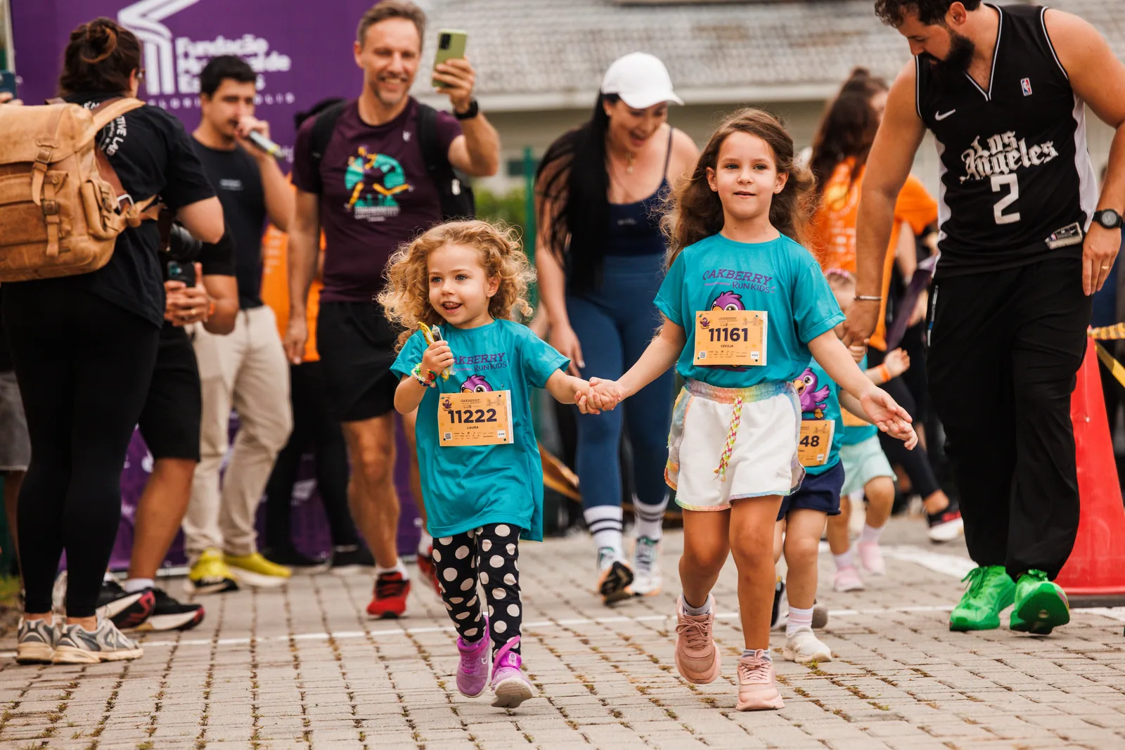 Duas meninas correm de mãos dadas na Oakberry Run Kids, com os pais filmando atrás