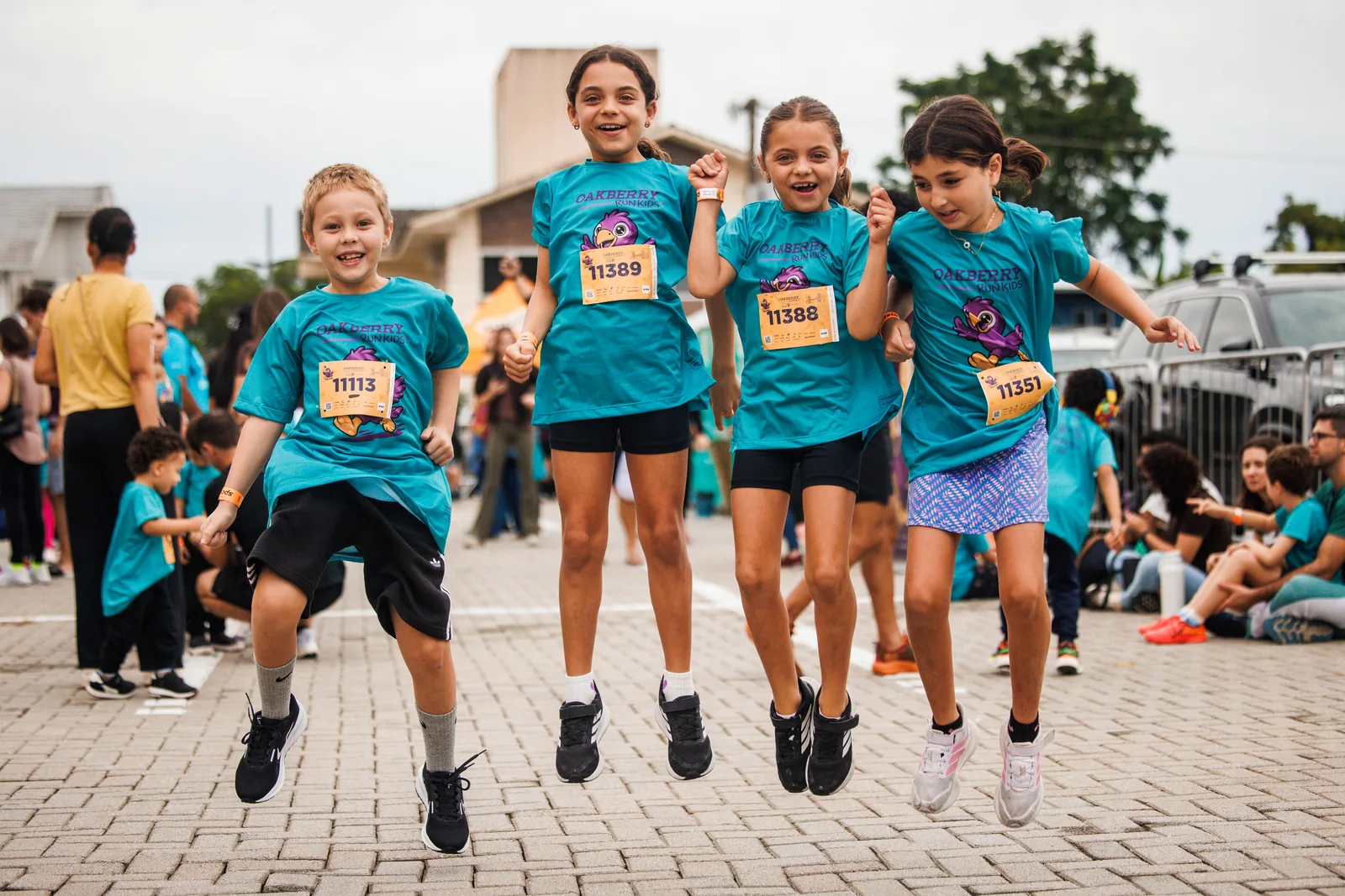 Quatro meninas de camiseta Oakberry Run Kids saltam juntas no calçamento