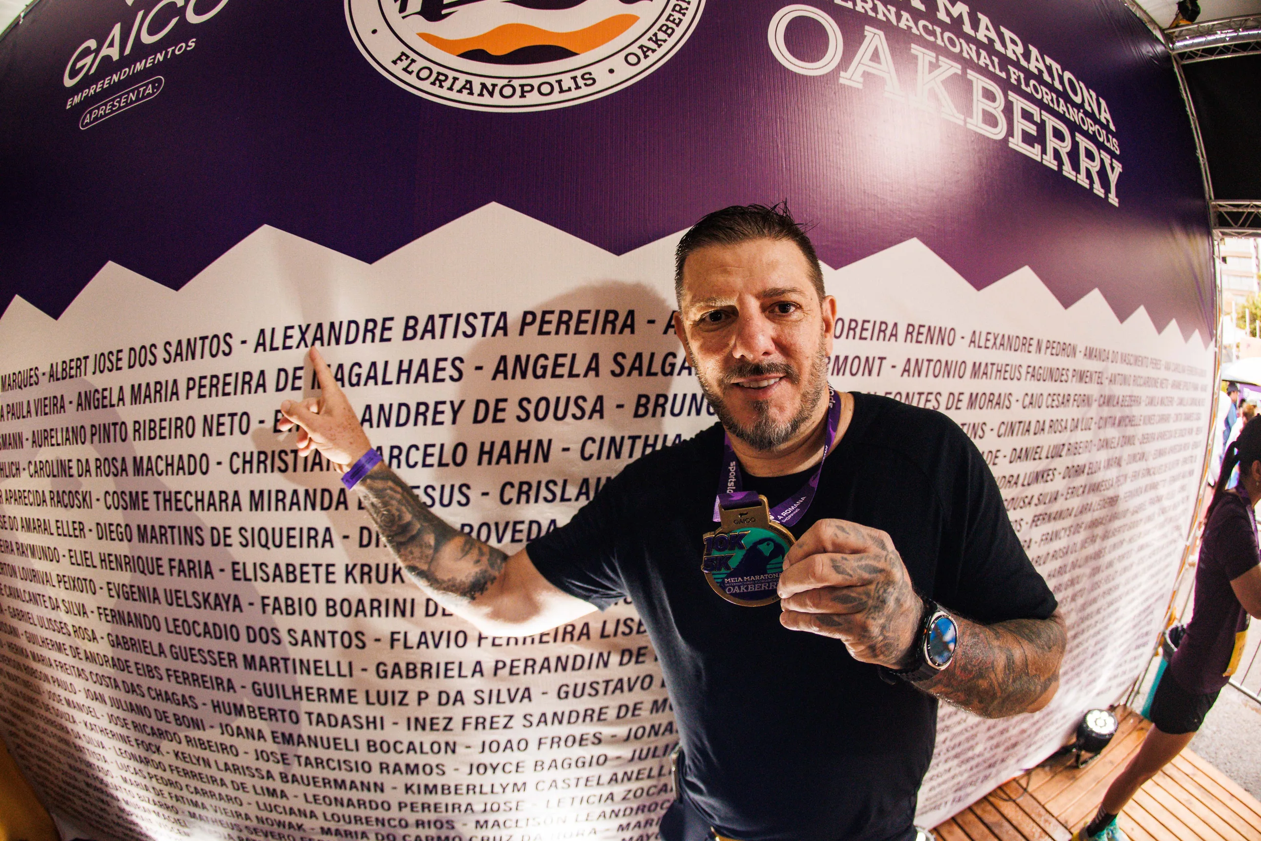 Atleta aponta o próprio nome no mural com os nomes dos inscritos e mostra a medalha