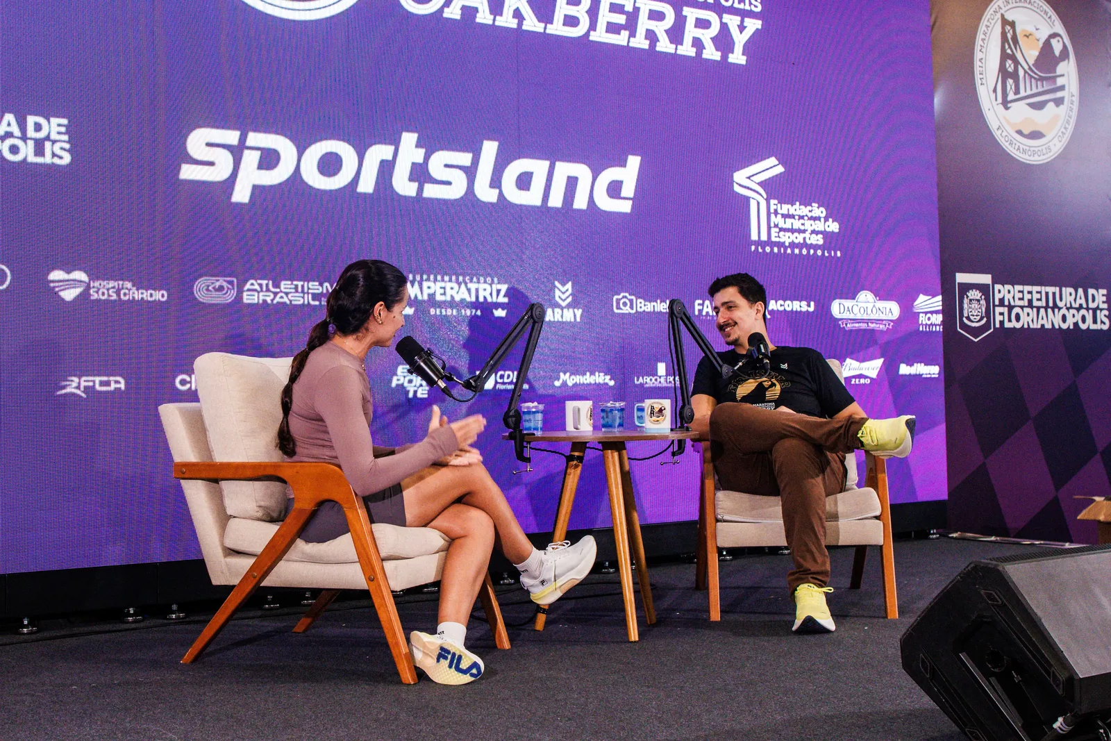 Gravação de podcast na Oakvillage: entrevistadora e convidado em poltronas diante do painel Sportsland