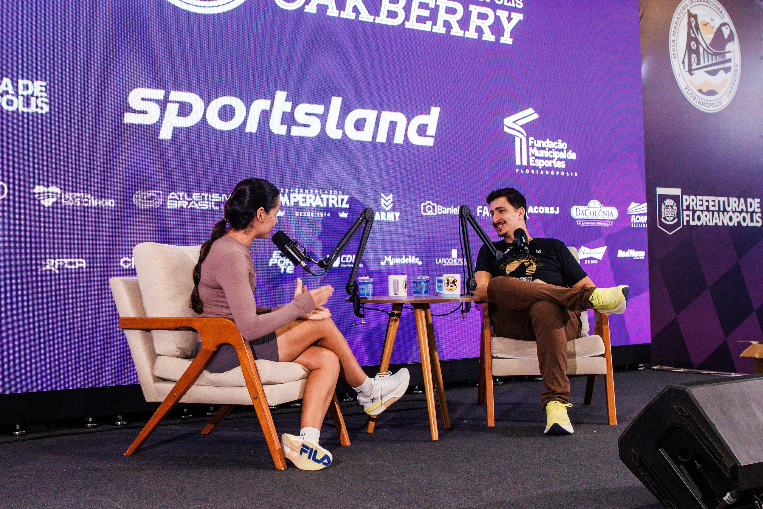 Gravação de podcast na Oakvillage: entrevistadora e convidado em poltronas diante do painel Sportsland
