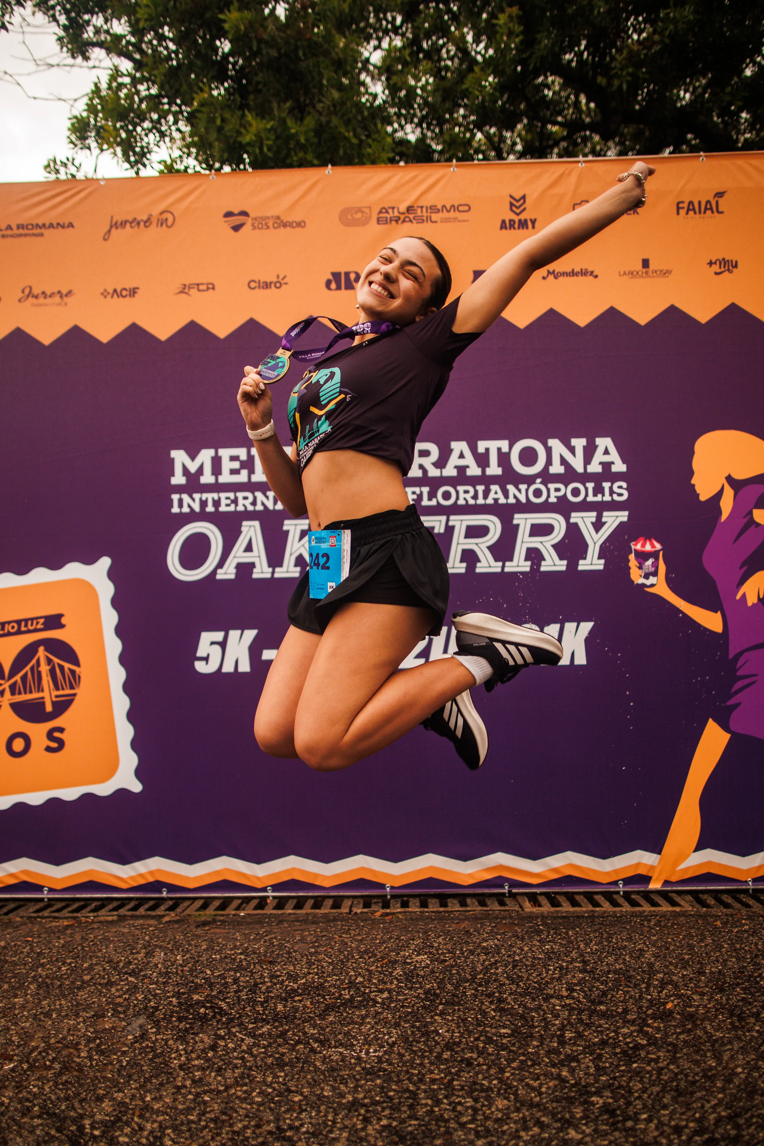 Corredora salta de braços abertos com a medalha diante do backdrop da Meia Maratona Oakberry