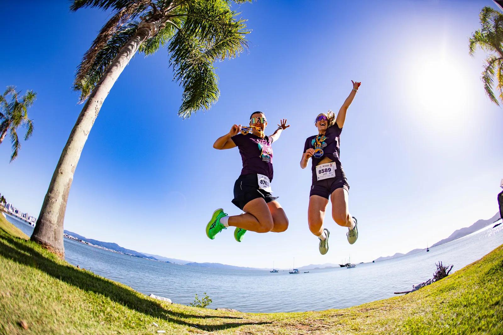 Duas corredoras saltando com as medalhas à beira da baía de Florianópolis, céu azul